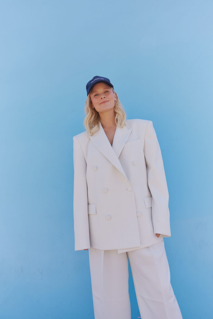 Blonde woman in navy blue hat and white suit, standing in front of a bright blue wall.